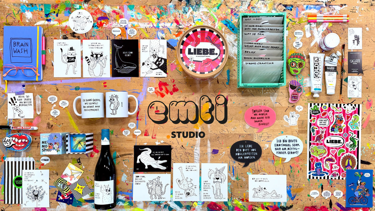 Tinis Arbeitstisch voller Farbspritzer, darauf Karten, Becher, Bubbles, Sticker, ein Puzzle und eine Weinflasche, in der Mitte der Schriftzug emti Studio