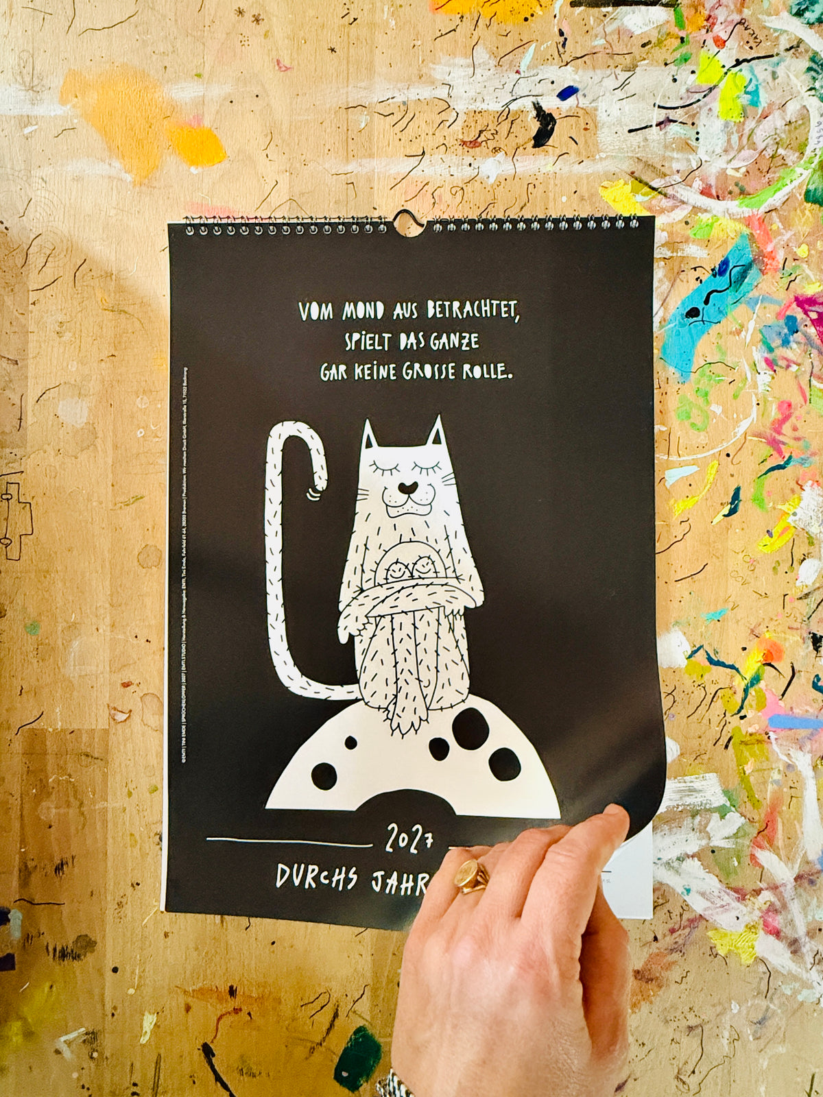 Schwarzes Deckblatt des Kalenders mit Spiralbindung: eine weiße Katze sitzt mit verschränkten Armen auf dem Mond, darüber „Vom Mond aus betrachtet, spielt das Ganze gar keine große Rolle.“