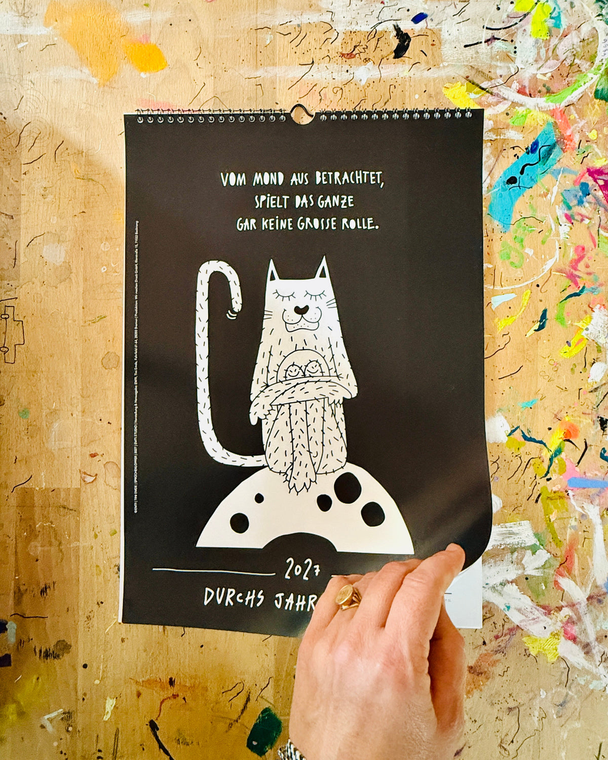 Schwarzes Deckblatt des Kalenders mit Spiralbindung: eine weiße Katze sitzt mit verschränkten Armen auf dem Mond, darüber „Vom Mond aus betrachtet, spielt das Ganze gar keine große Rolle.“