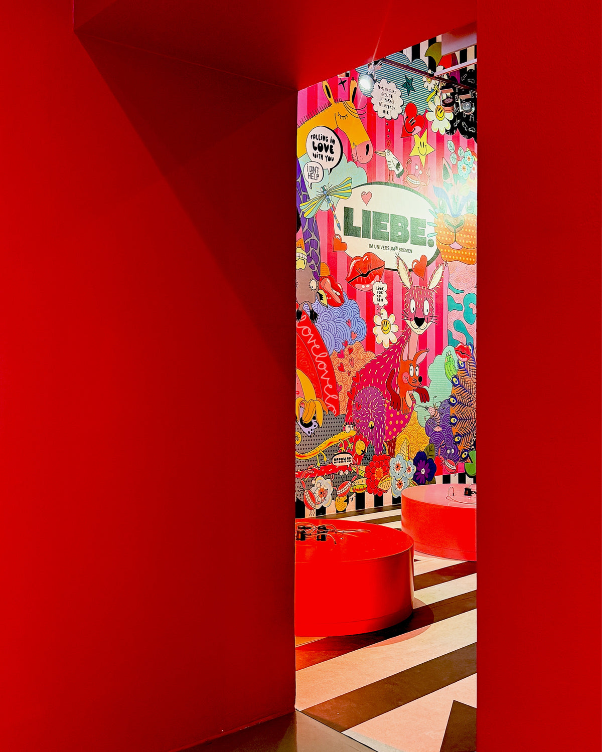Blick durch einen roten Durchgang auf die bunte Wandillustration mit dem Schriftzug „LIEBE.“ und zwei runde rote Podeste auf schwarz-weiß gestreiftem Boden