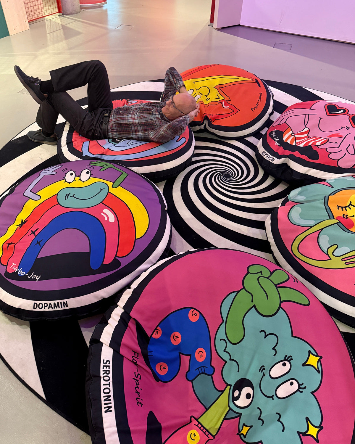Ein Mann liegt mit den Händen hinter dem Kopf auf runden Sitzkissen mit bunten Figuren, beschriftet mit „Dopamin“ und „Serotonin“, auf einem schwarz-weißen Spiralteppich