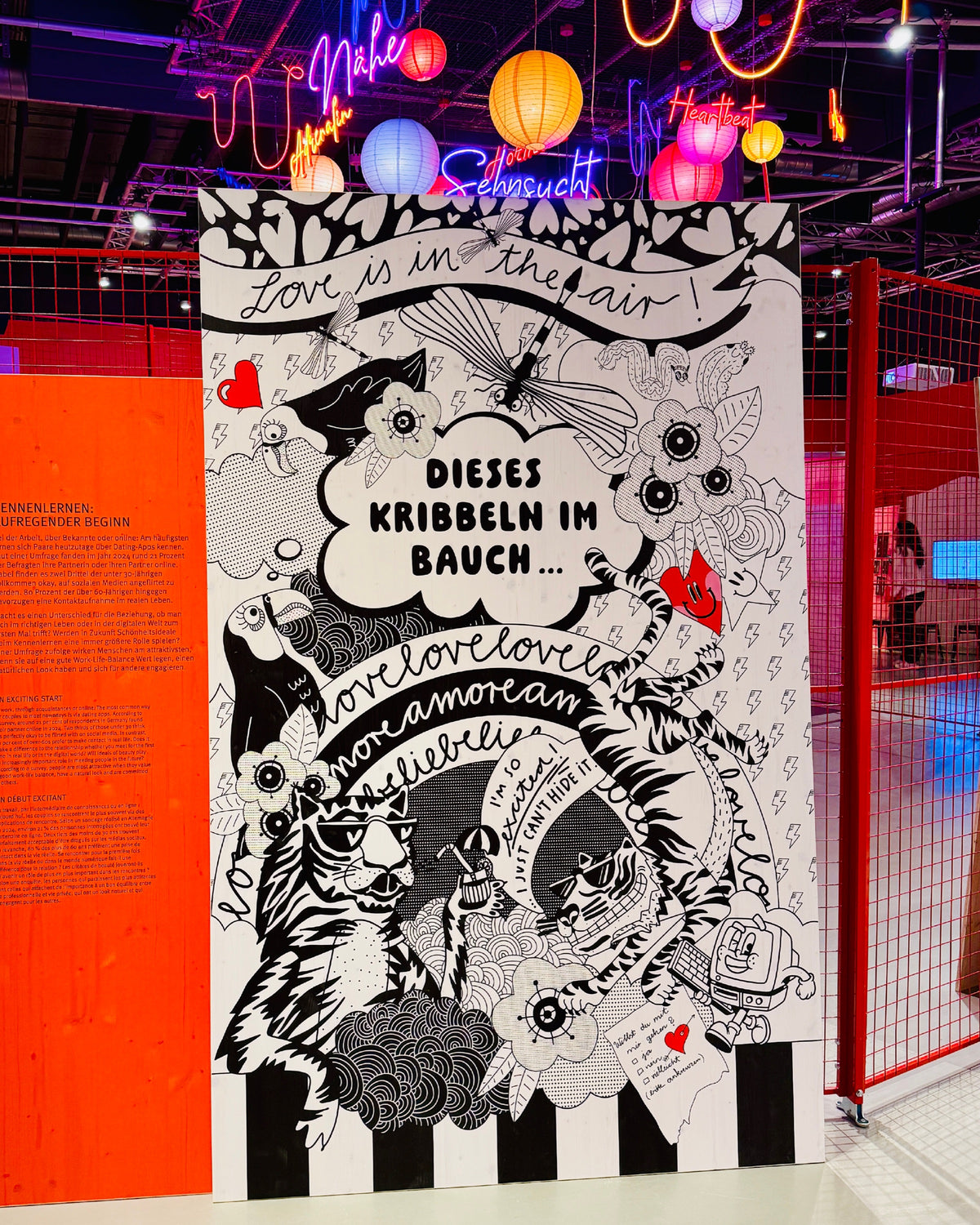 Schwarz-weiße Tafel mit den Schriftzügen „Love is in the air!“ und „Dieses Kribbeln im Bauch …“, darüber bunte Lampions und Neonschriften wie „Nähe“ und „Sehnsucht“