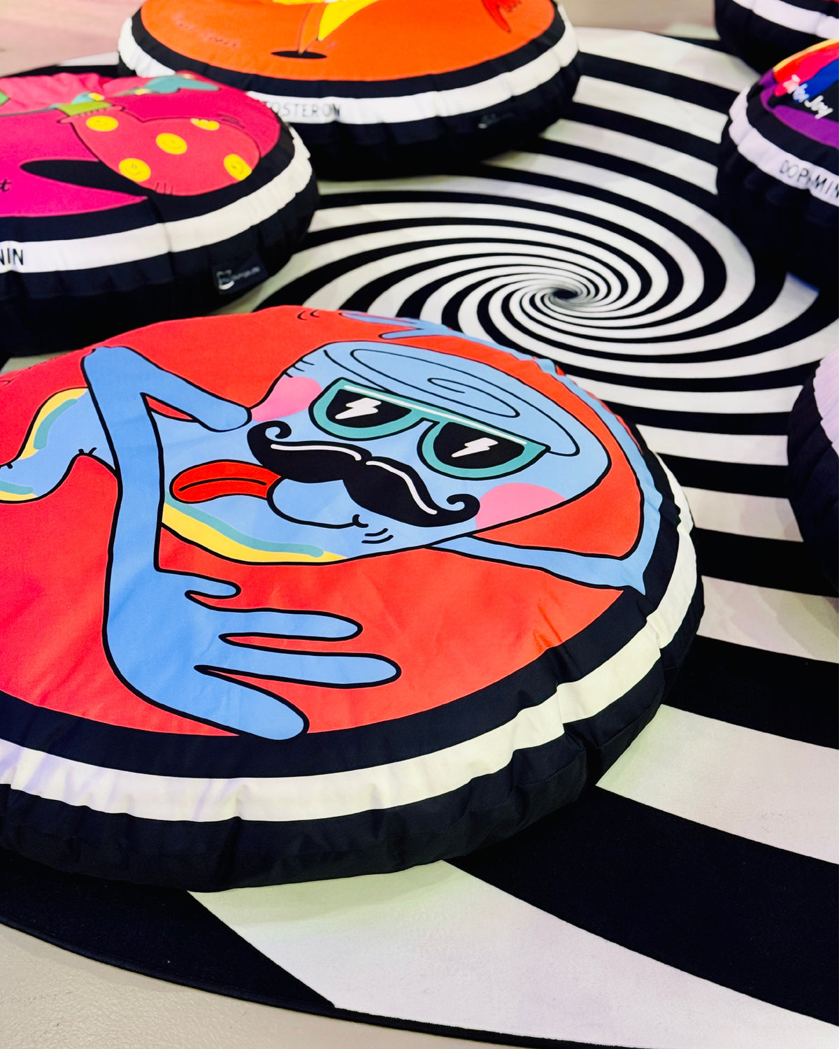 Nahaufnahme eines roten Sitzkissens mit einer blauen Figur mit Schnurrbart und Blitz-Sonnenbrille, dahinter weitere Kissen auf einem schwarz-weißen Spiralteppich