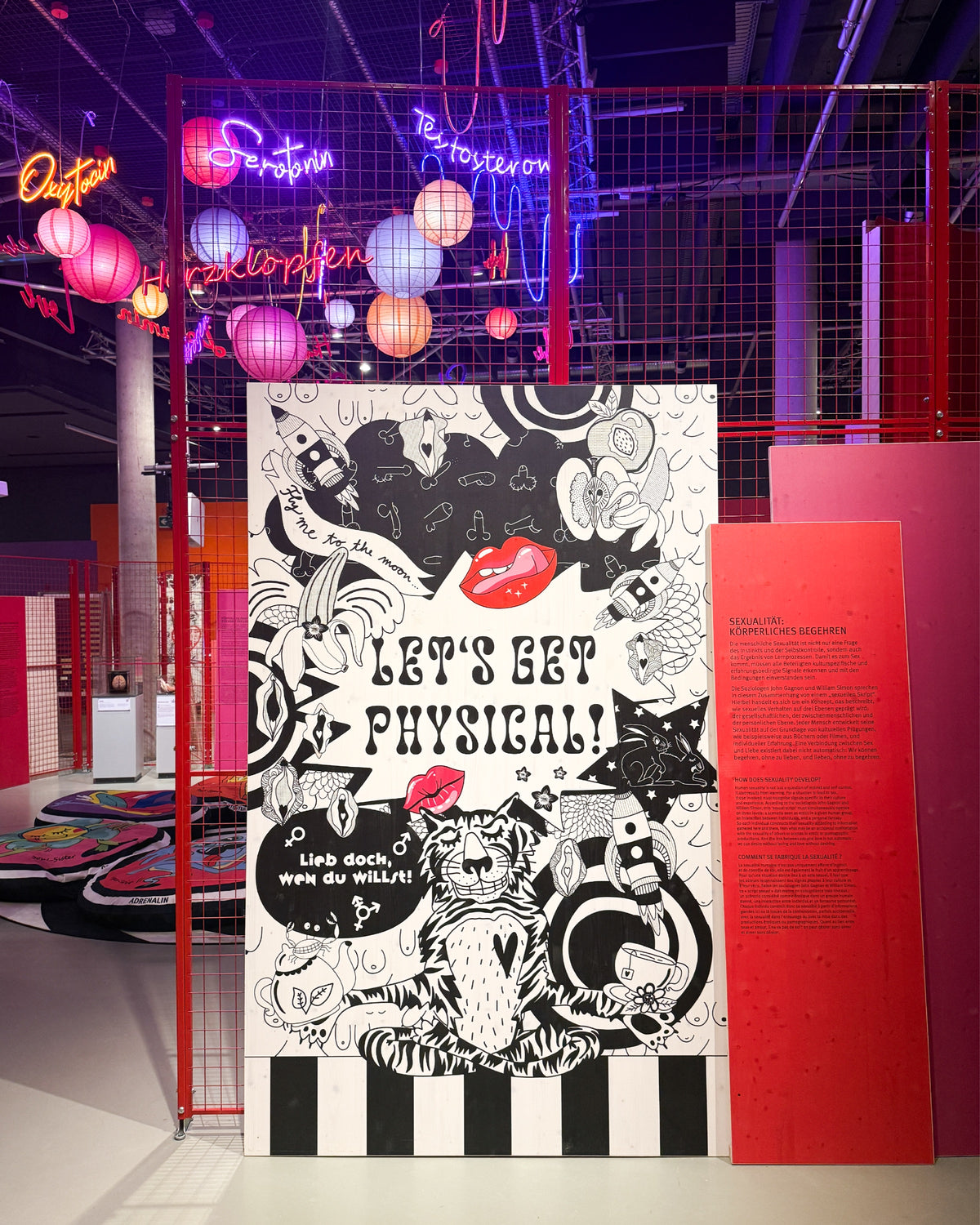 Schwarz-weiße Tafel mit den Schriftzügen „Let’s get physical!“ und „Lieb doch, wen du willst!“, einem Tiger und roten Lippen, darüber Lampions und Neonschriften wie „Serotonin“ und „Herzklopfen“