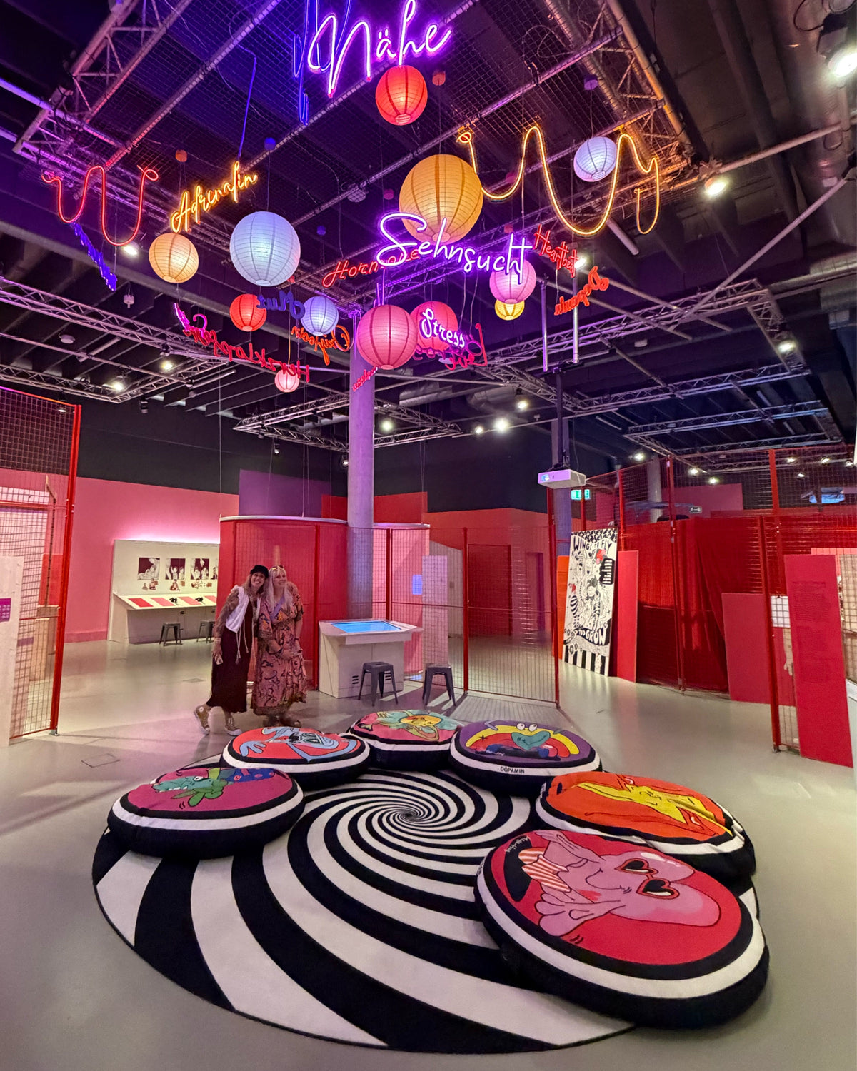 Blick in die Ausstellungshalle: bunte Lampions und Neonschriften wie „Nähe“ und „Sehnsucht“ unter der Decke, rote Gitterwände und runde Sitzkissen auf einem schwarz-weißen Spiralteppich