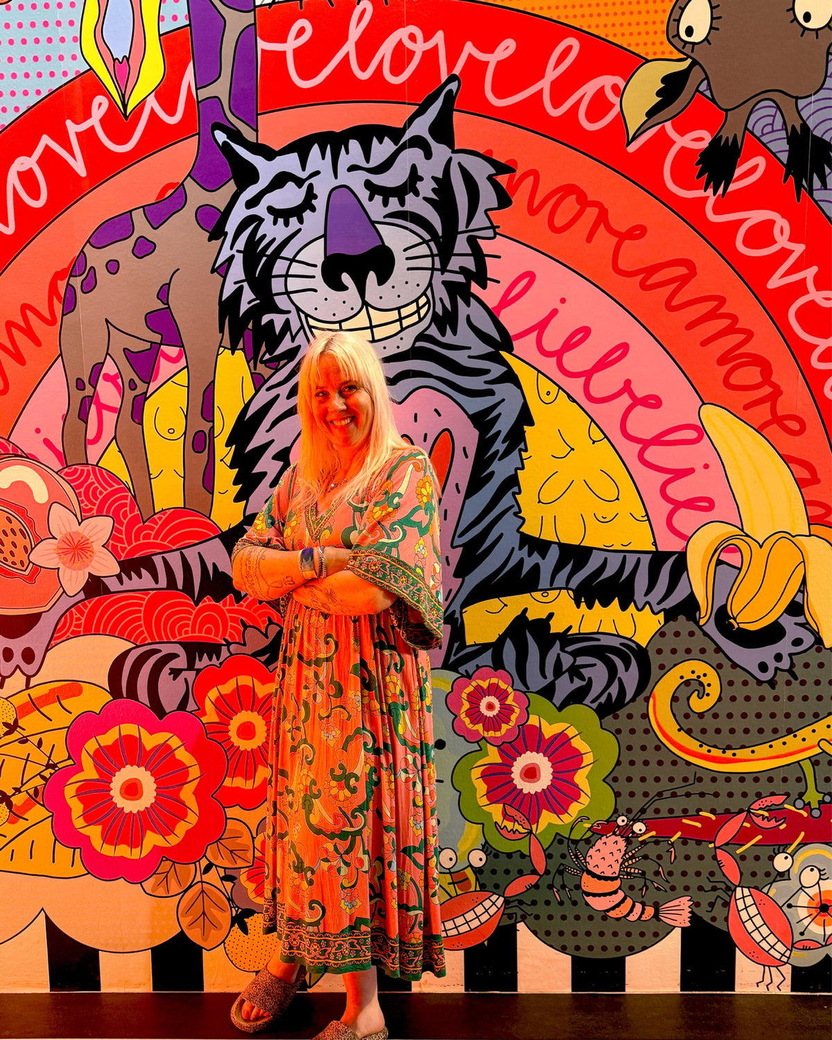 Eine Frau im geblümten Kleid steht lächelnd vor der Wandillustration mit einem grinsenden Tiger und dem Schriftzug „love love amore“ im Bogen