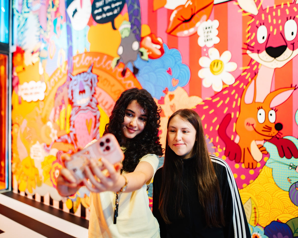 Zwei junge Frauen machen ein Selfie vor der Wandillustration mit Blumen, roten Lippen und bunten Tieren