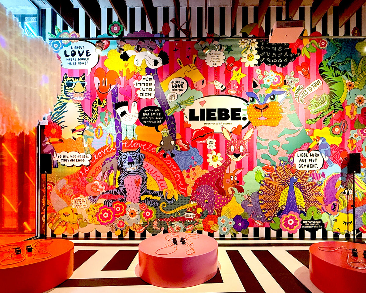 Die Wandillustration der Ausstellung „LIEBE.“ im Universum® Bremen: bunte Tiere, Blumen und Sprechblasen auf rosa Streifen um den Schriftzug „LIEBE.“, davor drei runde rote Podeste mit Telefonhörern