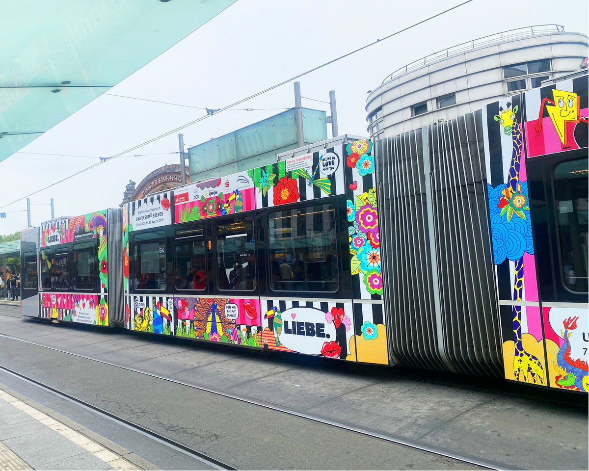 Die LOVETRAIN-Straßenbahn in Seitenansicht am Bremer Hauptbahnhof: Blumen, Giraffe, Wal und der Schriftzug „LIEBE.“, dahinter das Bahnhofsgebäude