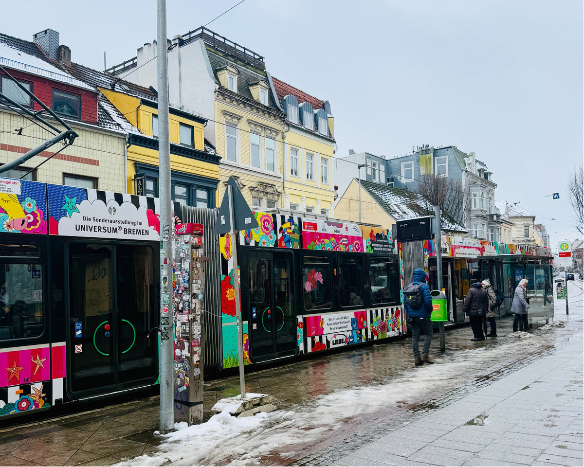 Die LOVETRAIN-Straßenbahn hält im Winter an einer Haltestelle vor bunten Altbauten, Fahrgäste warten auf dem verschneiten Gehweg