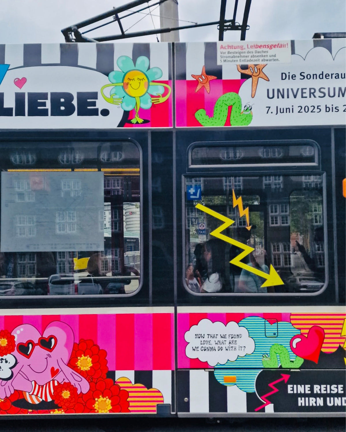 Nahaufnahme der LOVETRAIN-Straßenbahn: der Schriftzug „LIEBE.“ mit Smiley-Blume, ein gelber Blitz im Fenster und die Sprechblase „Now that we found love, what are we gonna do with it?“