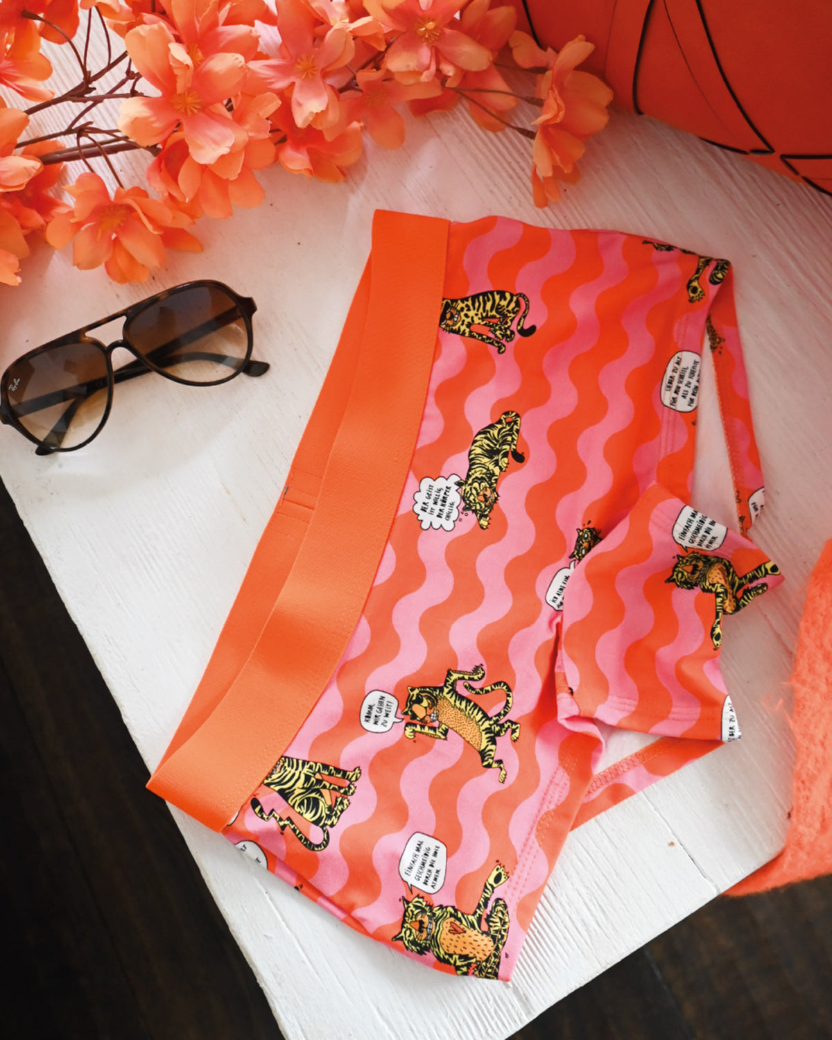 Schockhöschen mit Tigern und Sprechblasen auf rosa-orangen Wellen, daneben eine Sonnenbrille und orange Blüten