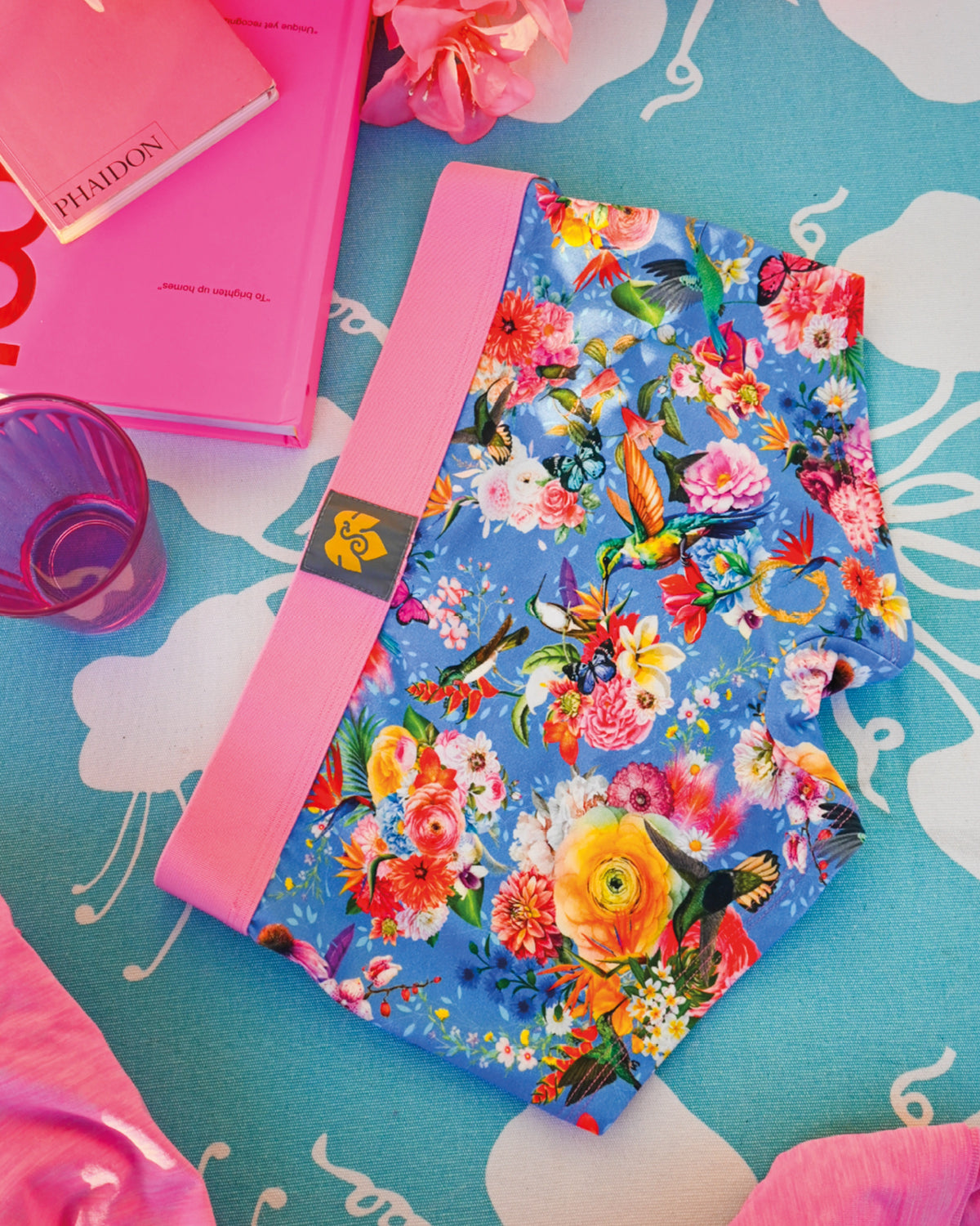 Schockhöschen mit Kolibris und Blumen auf blauem Grund, daneben rosa Bücher und ein Glas