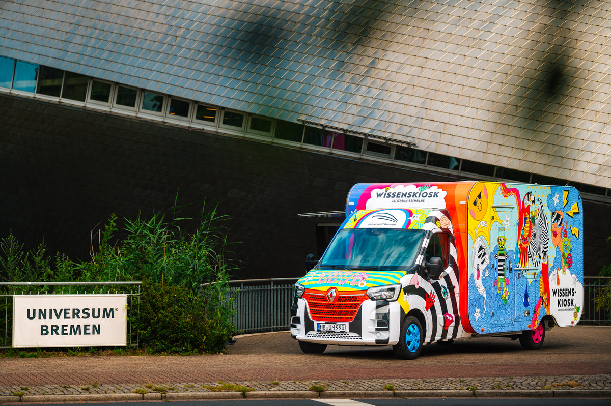Der Wissenskiosk – ein bunt folierter Transporter mit angehängtem, illustriertem Kiosk-Anhänger – steht vor dem Gebäude des Universum® Bremen, daneben ein Schild mit der Aufschrift „Universum Bremen“