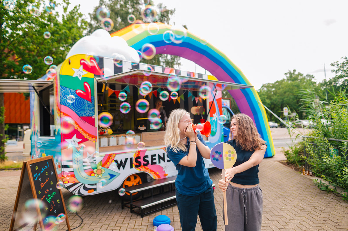 Zwei lachende Frauen pusten Seifenblasen vor dem Wissenskiosk mit aufblasbarem Regenbogen und Wolke
