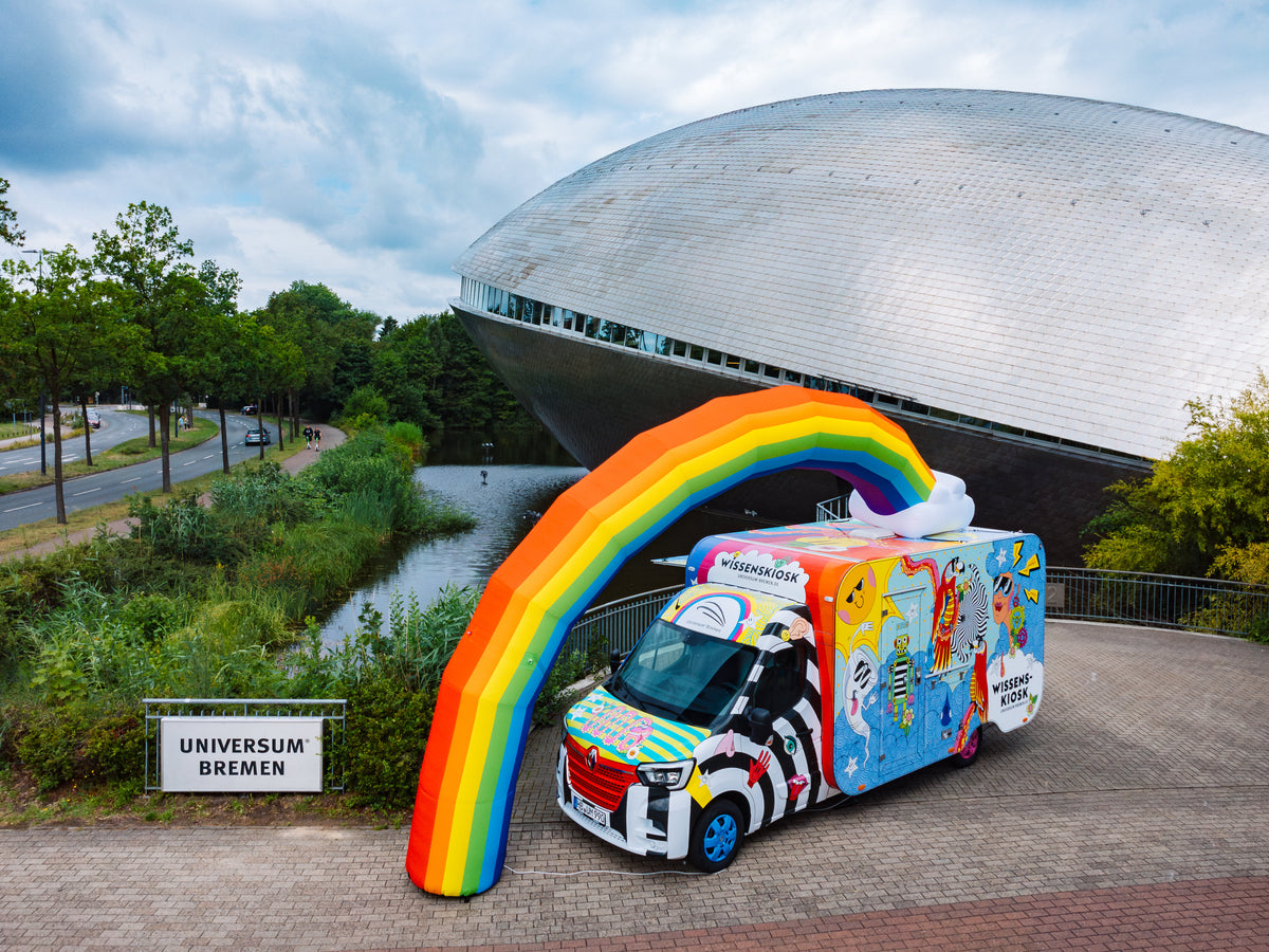 Luftaufnahme des Wissenskiosks aus buntem Transporter und Kiosk-Anhänger unter dem aufblasbaren Regenbogen vor der kuppelförmigen Architektur des Universum® Bremen