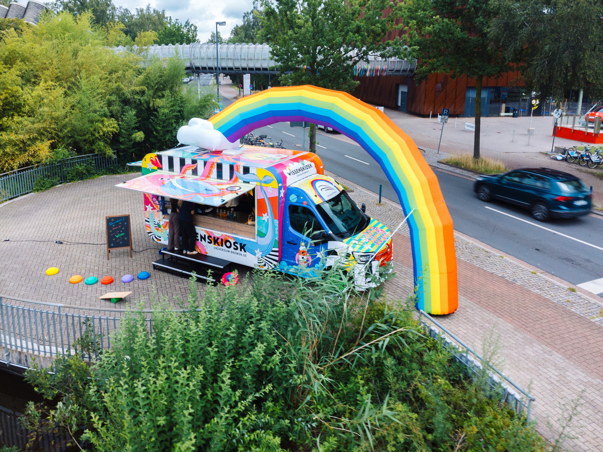 Luftaufnahme des geöffneten Wissenskiosks aus Transporter und Kiosk-Anhänger mit aufblasbarem Regenbogen auf einem Platz an einer Straße