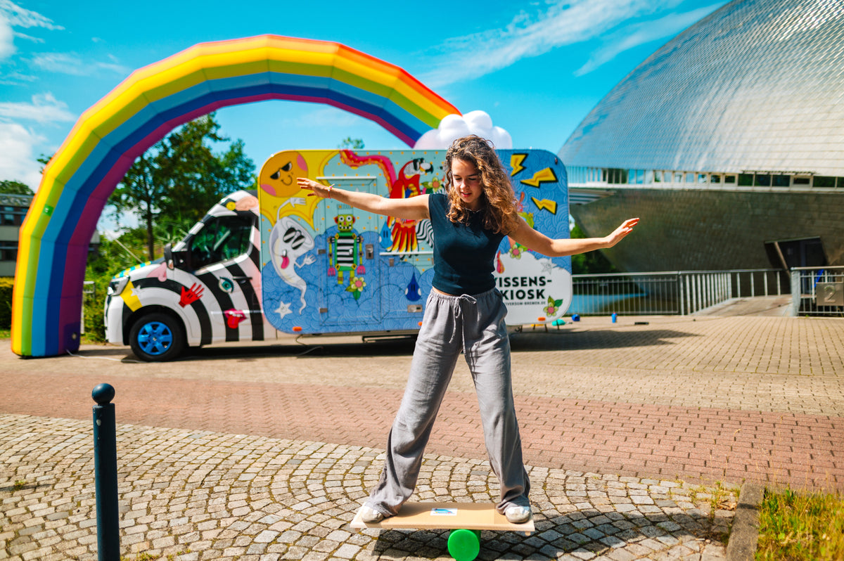 Eine Frau balanciert auf einem Wackelbrett vor dem Wissenskiosk mit Regenbogen und der Kuppel des Universum® Bremen im Hintergrund