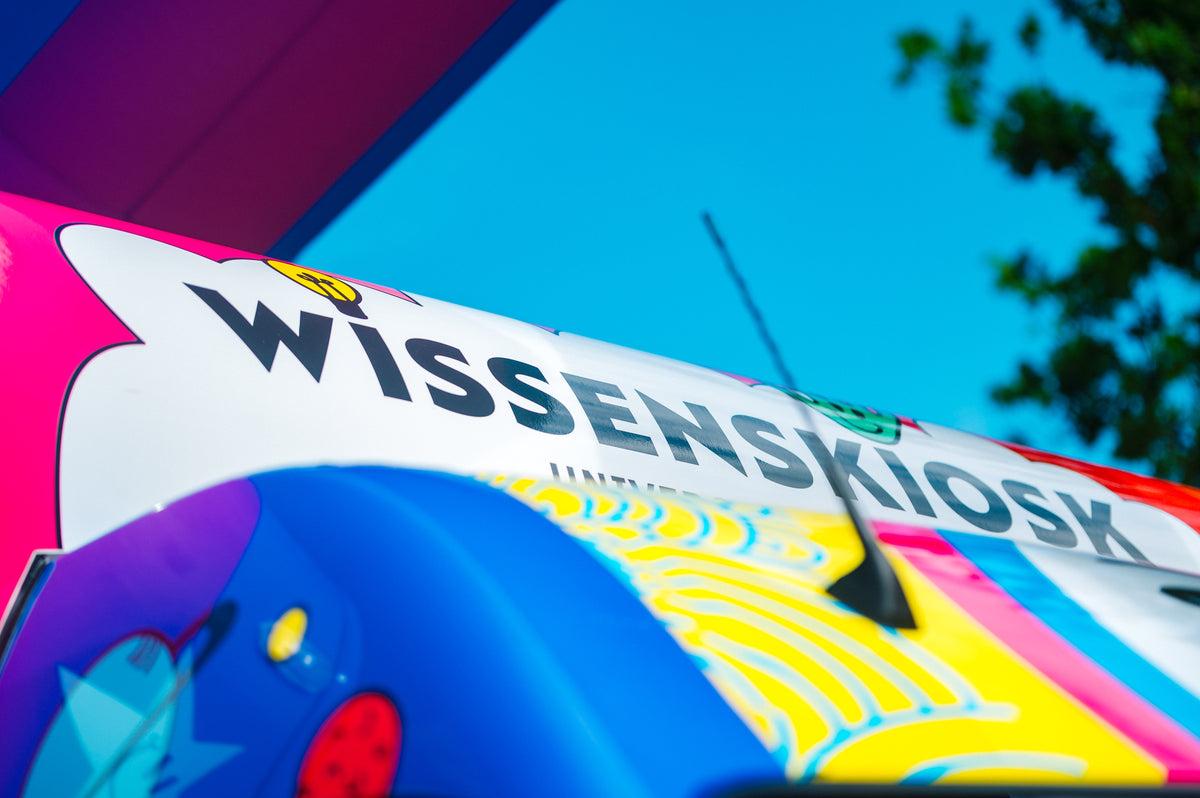 Nahaufnahme des „Wissenskiosk“-Schriftzugs auf dem bunt folierten Anhängerdach vor blauem Himmel
