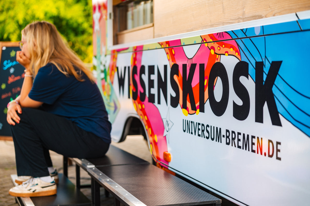 Eine Frau sitzt auf einer Bank neben dem Wissenskiosk, daneben groß der Schriftzug „Wissenskiosk – universum-bremen.de“