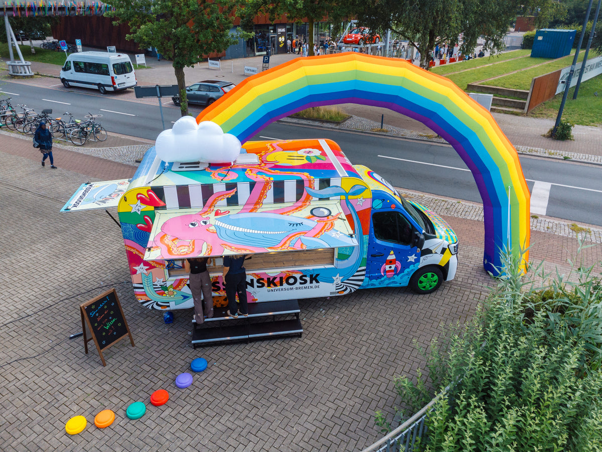 Luftaufnahme des geöffneten Wissenskiosks mit Regenbogen, davor bunte Sitzhocker; zwei Personen am Tresen