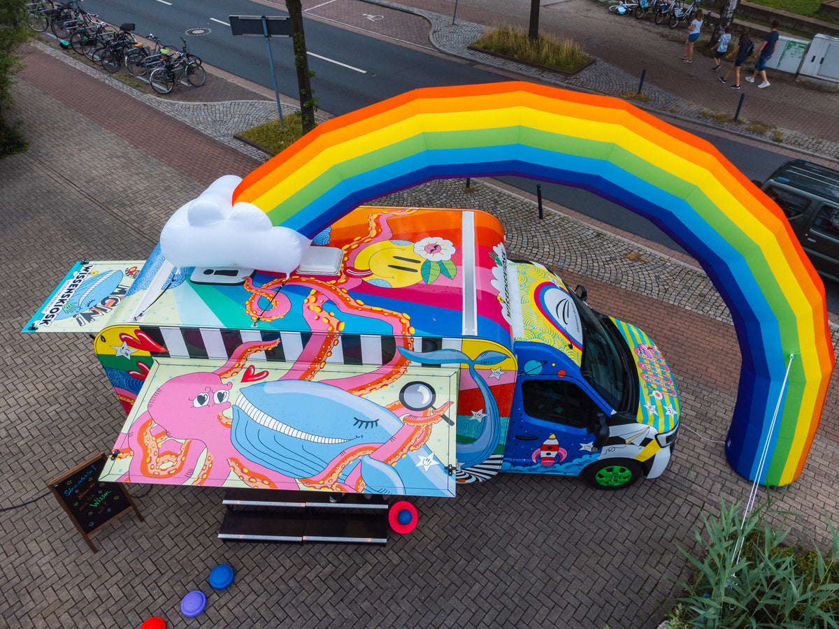 Schrägansicht von oben auf den Wissenskiosk aus vollflächig bunt illustriertem Transporter und Kiosk-Anhänger mit aufblasbarem Regenbogen und Wolke