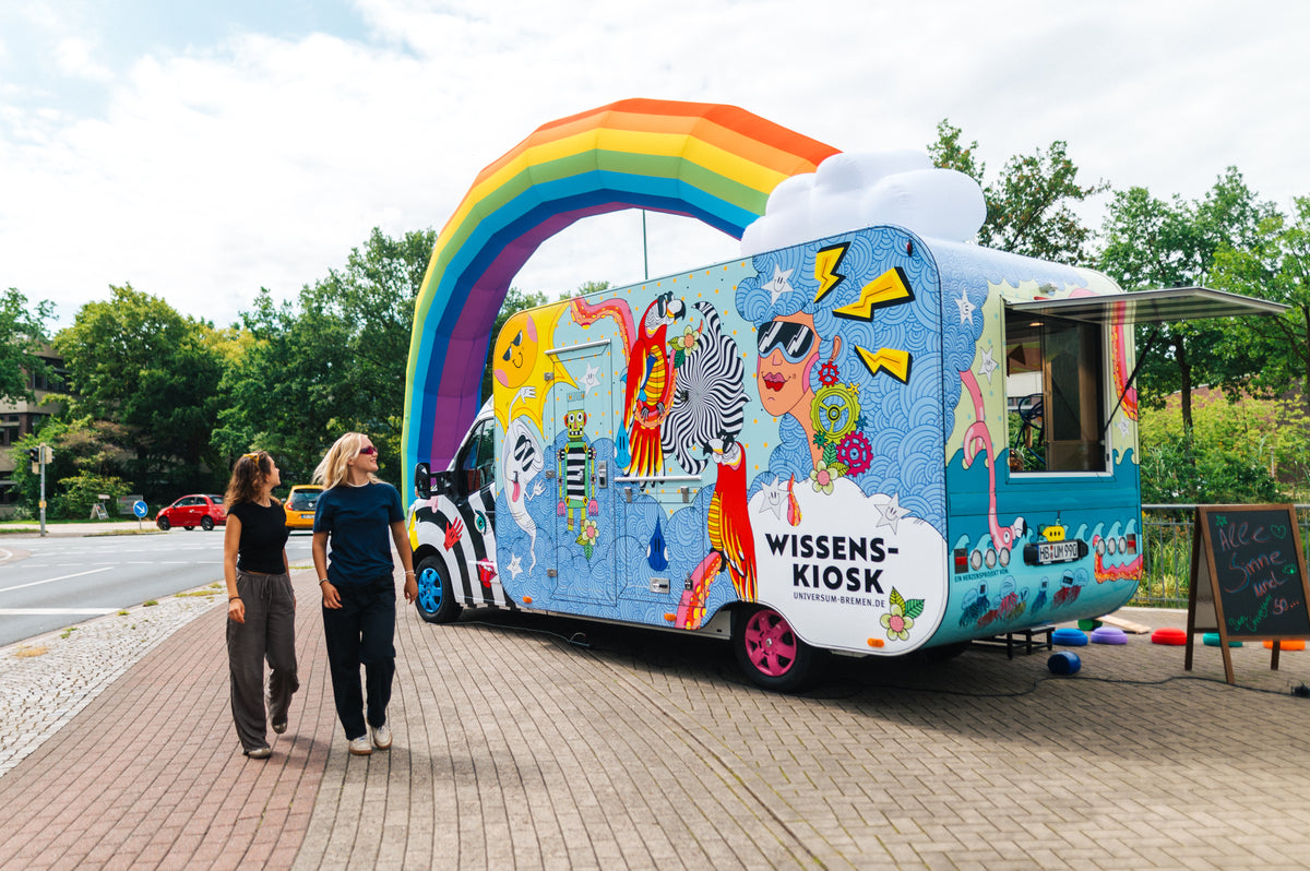 Zwei Frauen gehen am Wissenskiosk vorbei – einem bunt folierten Transporter mit Kiosk-Anhänger unter dem aufblasbaren Regenbogen