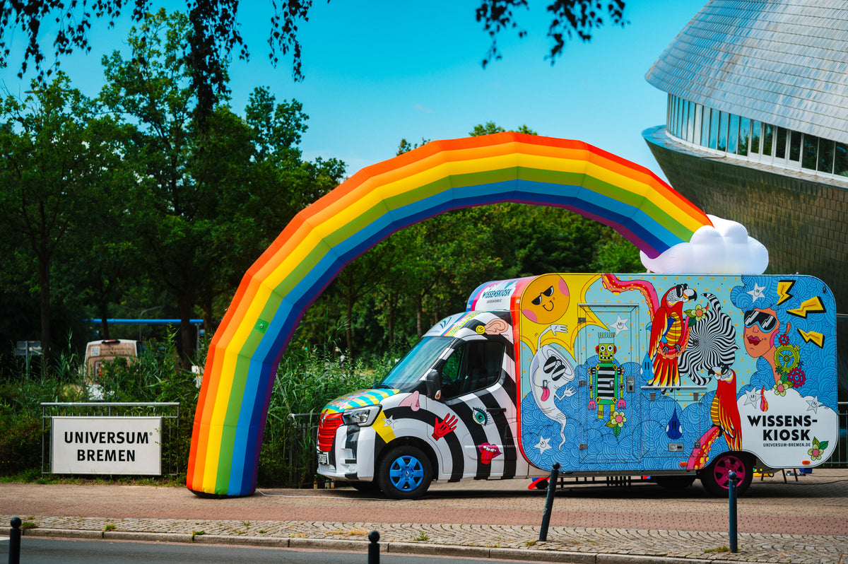 Der Wissenskiosk aus buntem Transporter und Kiosk-Anhänger mit aufblasbarem Regenbogen am Eingang des Universum® Bremen, daneben das Universum-Schild