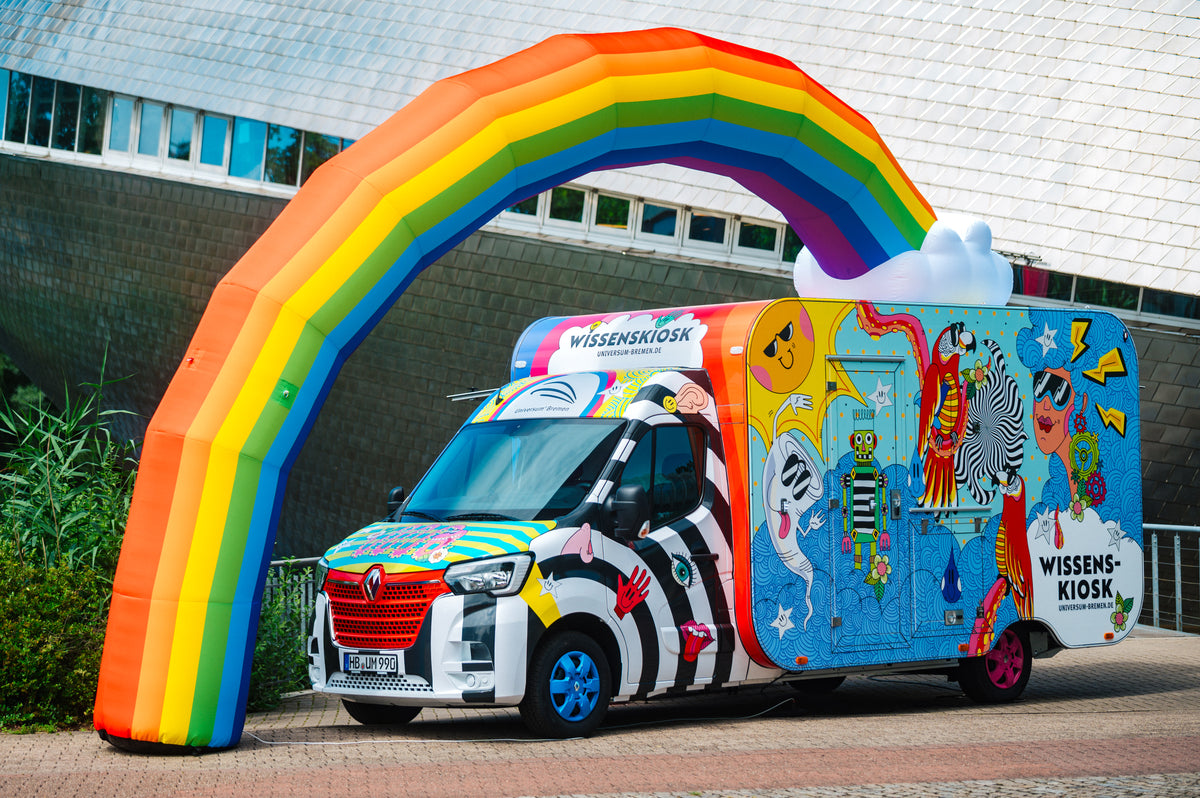 Frontansicht des Wissenskiosks: bunt illustrierter Transporter mit angehängtem Kiosk-Anhänger unter dem aufblasbaren Regenbogen vor dem Universum® Bremen