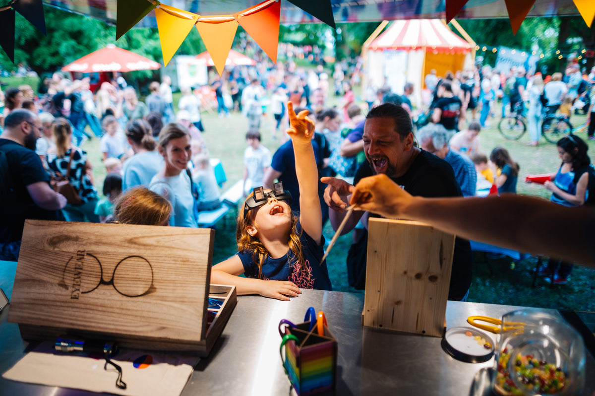 Ein lachendes Kind mit Papp-Brille zeigt nach oben, neben ihm ein Mann mit offenem Mund, am Experimentier-Stand mit Holzkästen auf einem Festival mit Wimpelketten